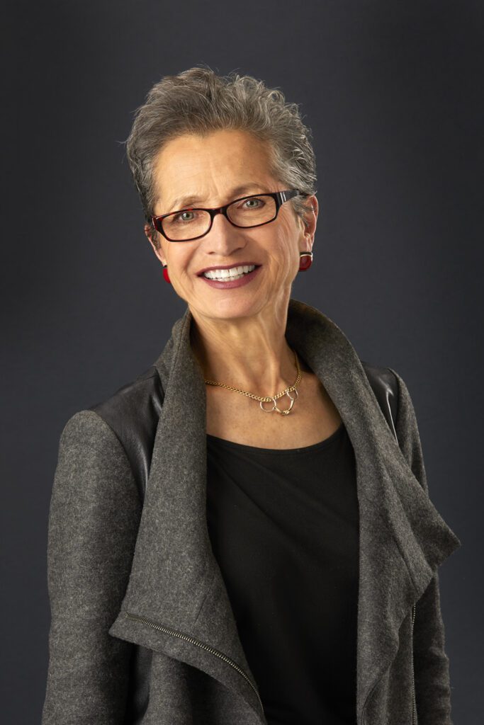 A woman with short hair and glasses wearing a gray jacket.