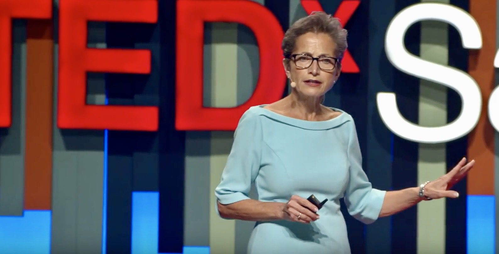 A woman in blue dress standing on stage with red ted logo.